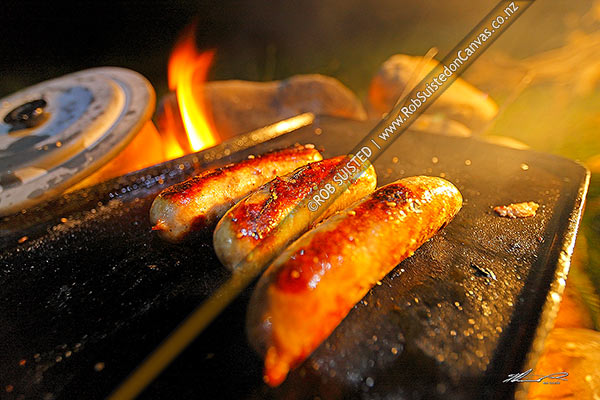 Photo of Sausages cooking and barbecuing on open campfire and hotplate at a night time campsite.,, New Zealand (NZ)